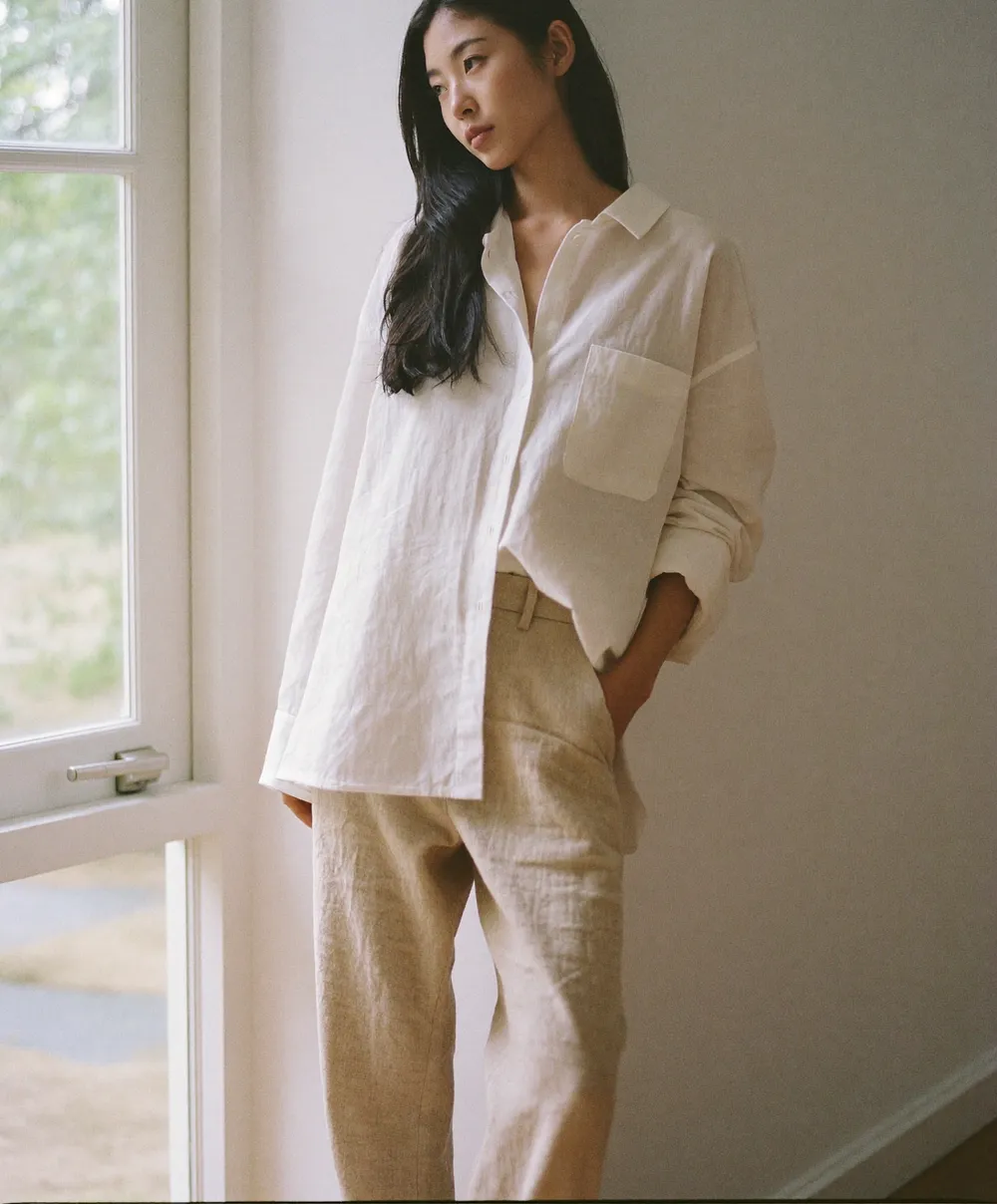 창가의 자연광 아래 화이트 리넨 셔츠와 베이지 리넨 팬츠를 입은 모델의 룩북 컷